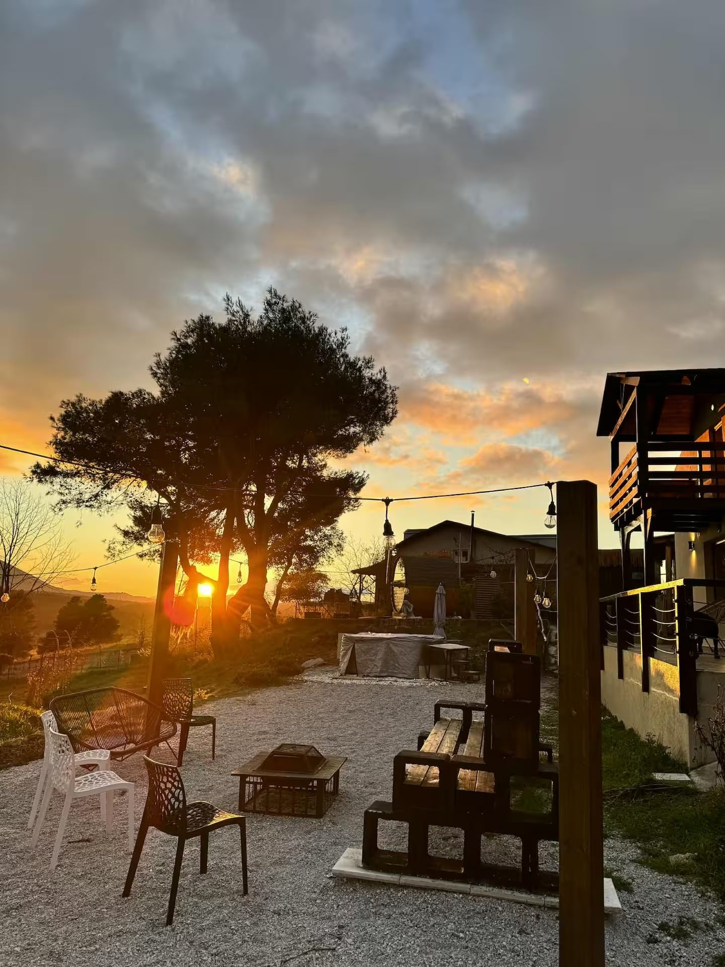 Sunset behind a pine tree, with the fire pit, garden chairs and string lights beside the house