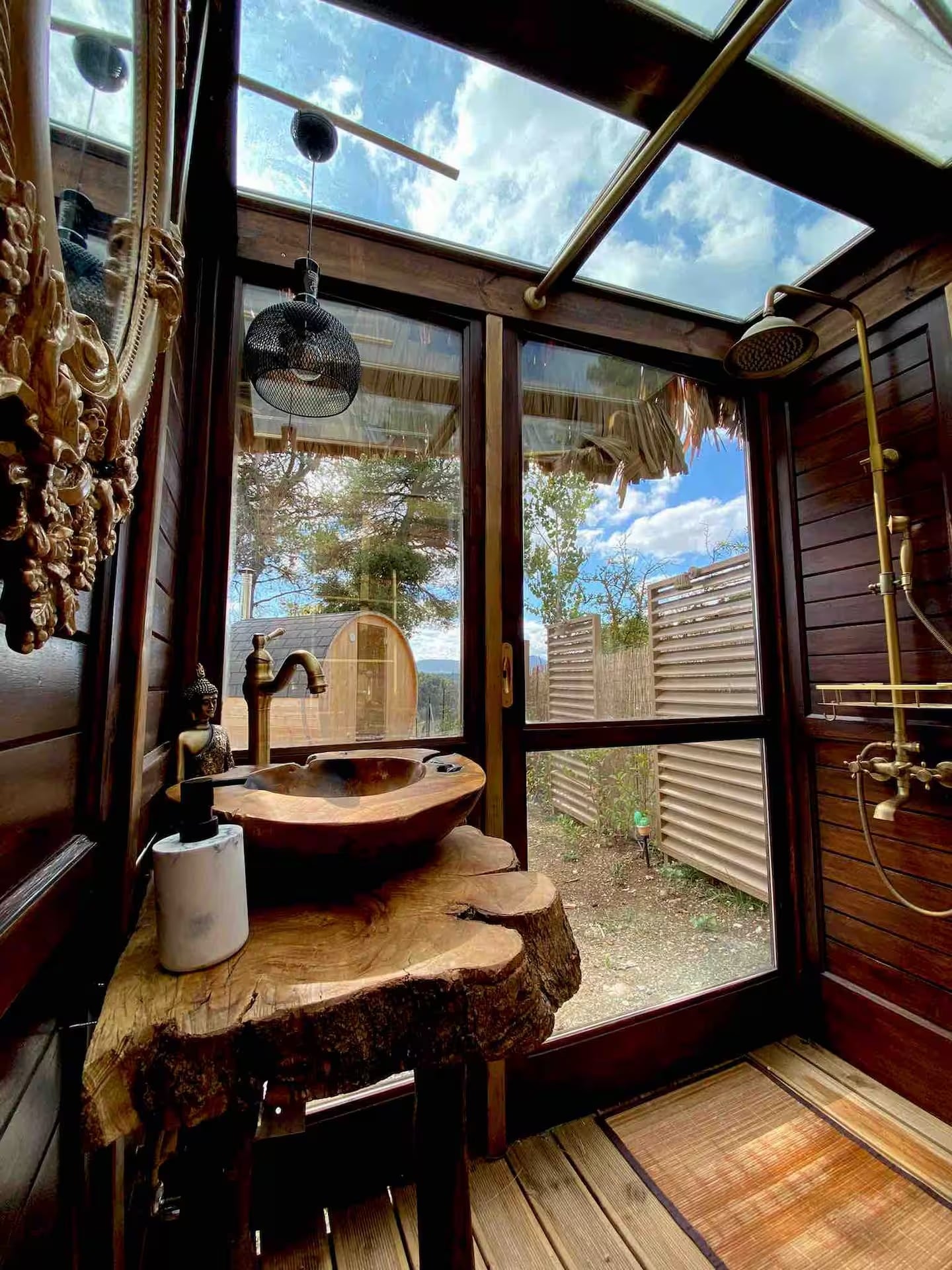 The outdoor wooden bathroom with a glass roof, brass shower and a wooden basin on a live-edge slab